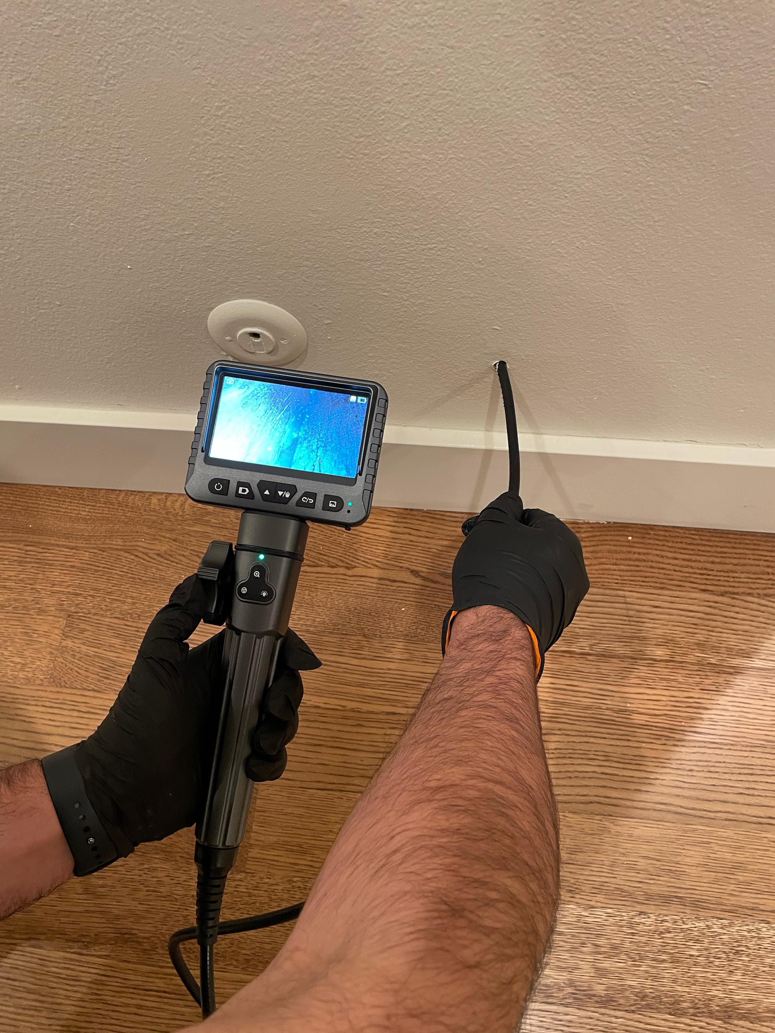 Inspector wearing black gloves threading borescope cable into wall through a small hole while viewing the LCD screen