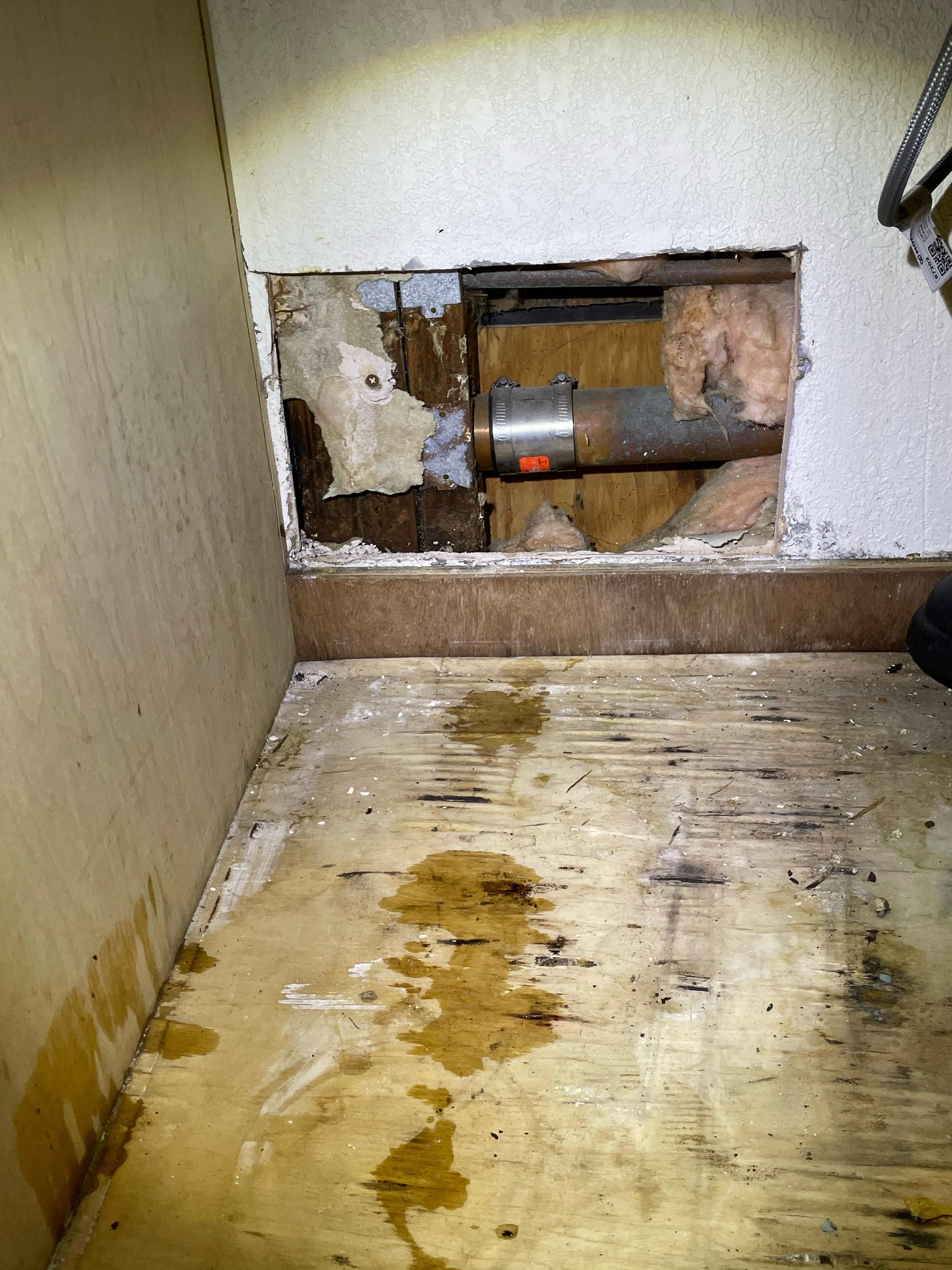 Opened wall section revealing water damage, rotted wood, ductwork, and stained subfloor behind drywall